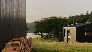 A rustic cabin with a metal roof and a chimney, surrounded by a grassy area and trees, with a body of water in the background. - Raus | Cabins am See mit Sauna + Hot-Tub (Lenzen)