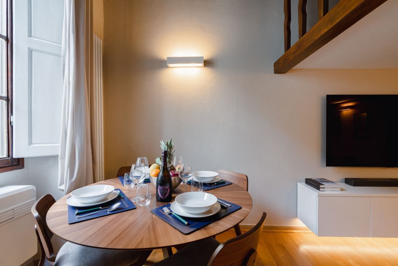 A dining area with a round table, chairs, a wall-mounted light, a TV, and a white cabinet.