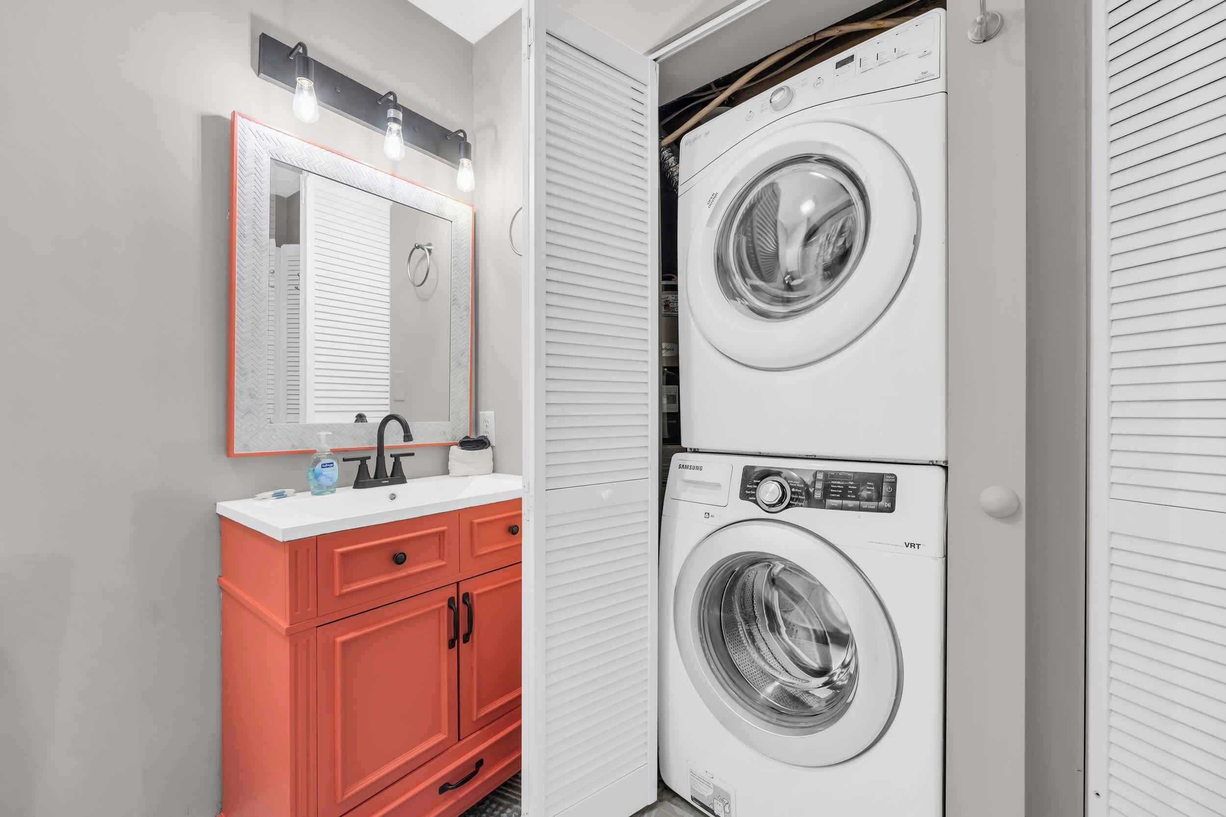 A laundry room with two stacked washing machines, a sink with a mirror, and a red cabinet.