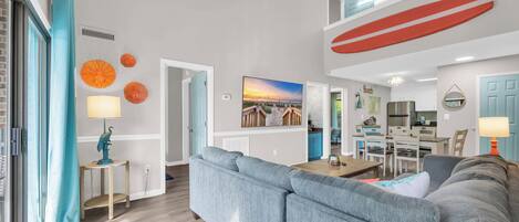 A modern living room with a grey sectional sofa, a dining area with a wooden table, and a kitchen in the background.