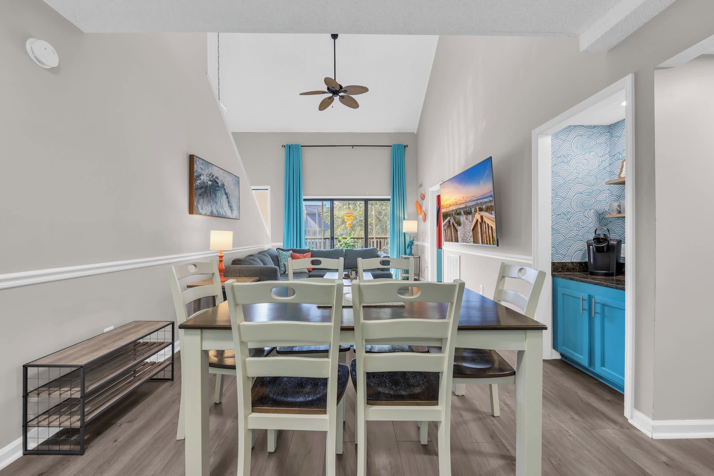 A dining area with a high ceiling, a ceiling fan, and a view of the living room through a window with blue curtains.