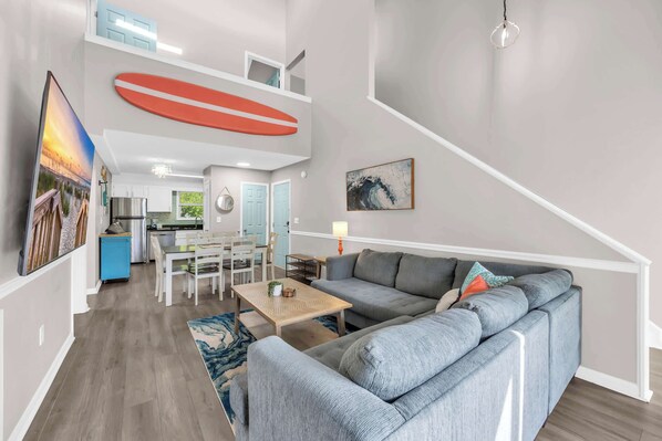 Een moderne woonkamer met een grijze hoekbank, een houten salontafel en een blauw vloerkleed. In de achtergrond is een keuken met witte kasten en een eethoek met een witte tafel en stoelen te zien.