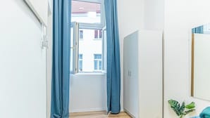 Ein Schlafzimmer mit Fenster, Vorhängen, einem Bett, einem Kleiderschrank und einer kleinen Pflanze.