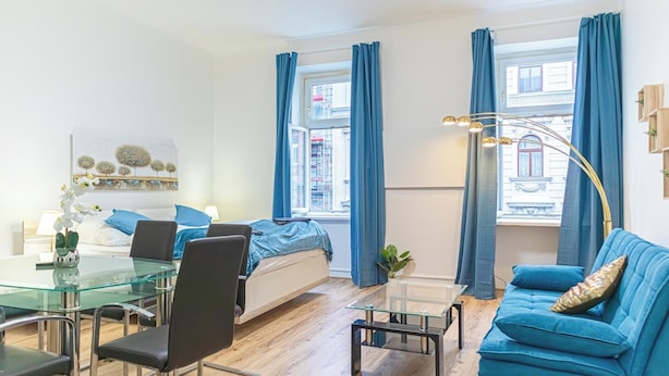 Ein modernes Zimmer mit einem Glastisch, einem blauen Sofa, einem Bett mit blauer Bettdecke und großen Fenstern mit blauen Vorhängen.