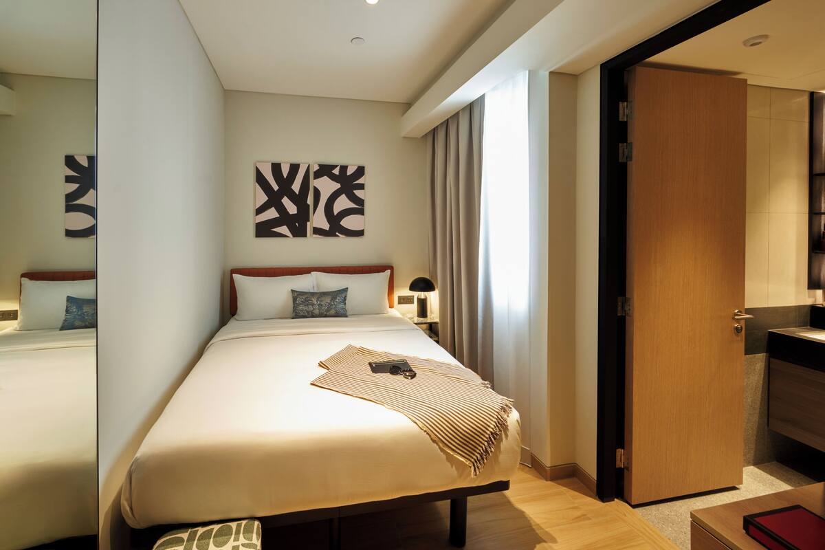 Habitación de hotel con dos camas, una mesita de noche con lámpara, un espejo grande y arte abstracto en la pared.