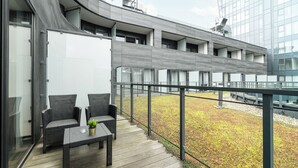 Varanda moderna ao ar livre com móveis de vime e uma pequena mesa, com vista para um jardim paisagístico e um edifício com grandes janelas.