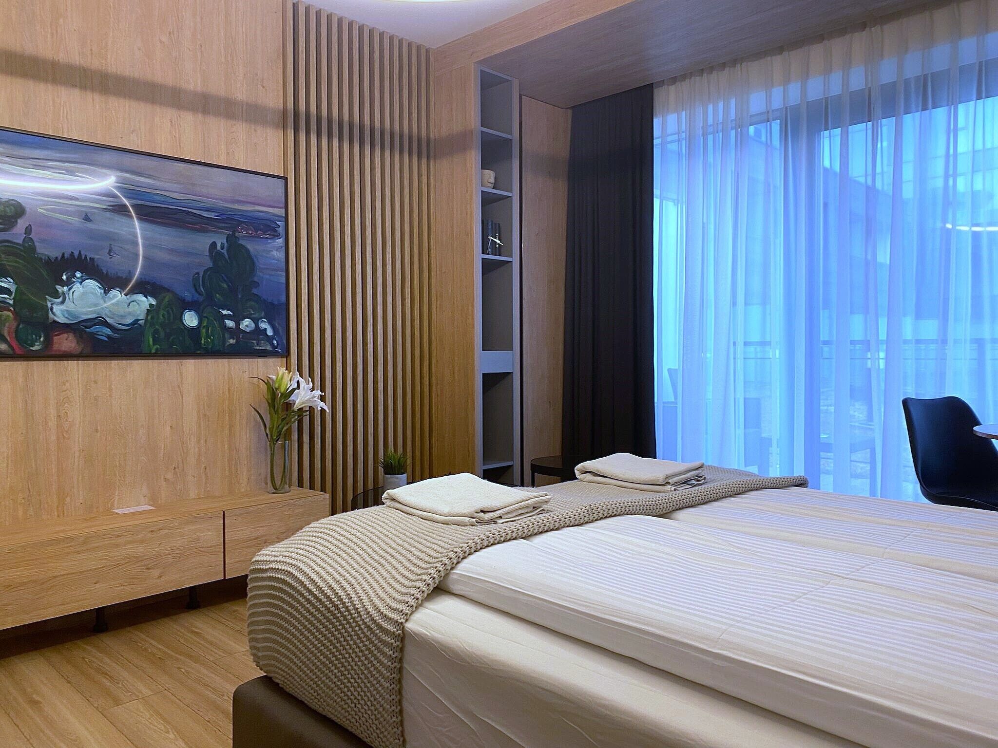 A modern hotel room with a large bed, a wooden panel wall, a painting, a vase with flowers, and a view of the city through sheer curtains.
