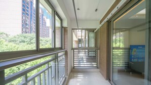 A corridor with a glass door leading to a room, a balcony with a railing, and a view of tall buildings and greenery outside.