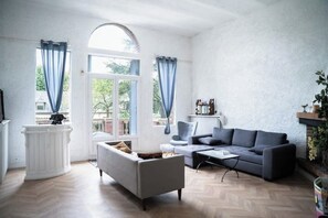 A living room with a grey sofa, a white armchair, a coffee table, and a white console table. There are two windows with blue curtains, and a fireplace with a mantel. - Large 10-bedroom manor house, sleeps up to 20—view of the Seine, Les Andelys, 1 hour from Paris (Les Andelys)