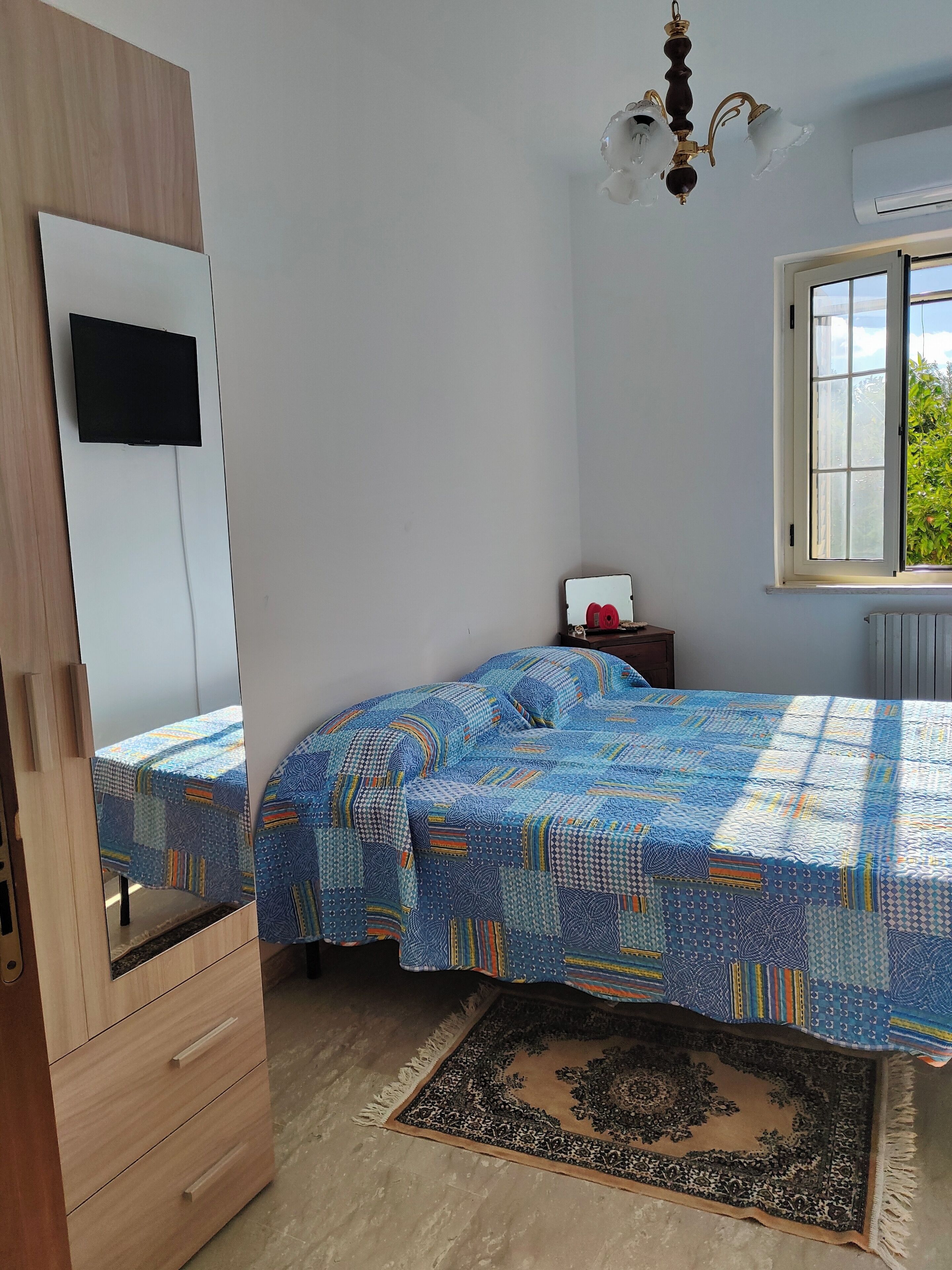 Une chambre à coucher avec un lit recouvert d’une couverture à motifs, une armoire en bois, une petite table de chevet avec une lampe, une fenêtre donnant sur la verdure et un luminaire au plafond.