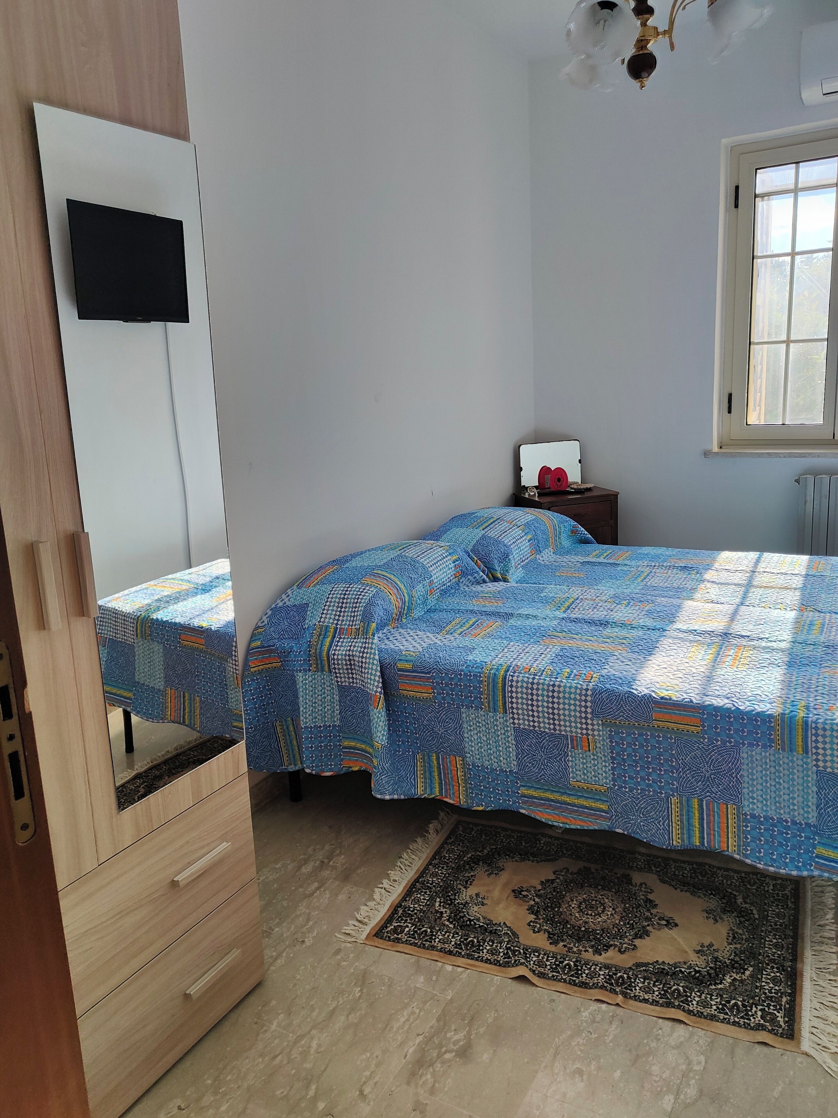 A bedroom with a bed covered in a patterned bedspread, a wooden wardrobe, a small bedside table with a lamp, and a window with a view of trees.