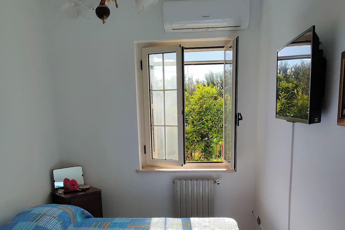 A bedroom with a bed, a window with a view, an air conditioner, and a mirror on the wall.