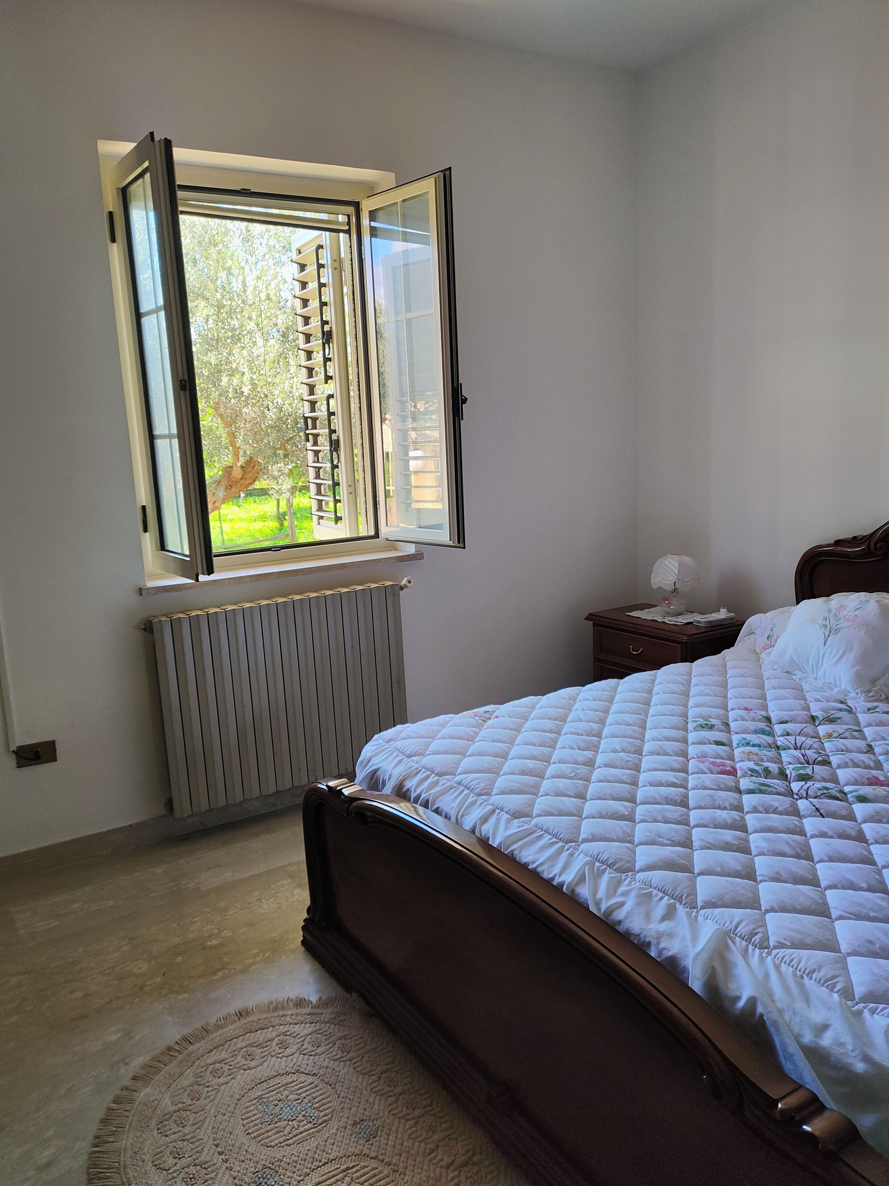 Un dormitorio con cama, ventana con vista a follaje, radiador y mesita de noche con lámpara.
