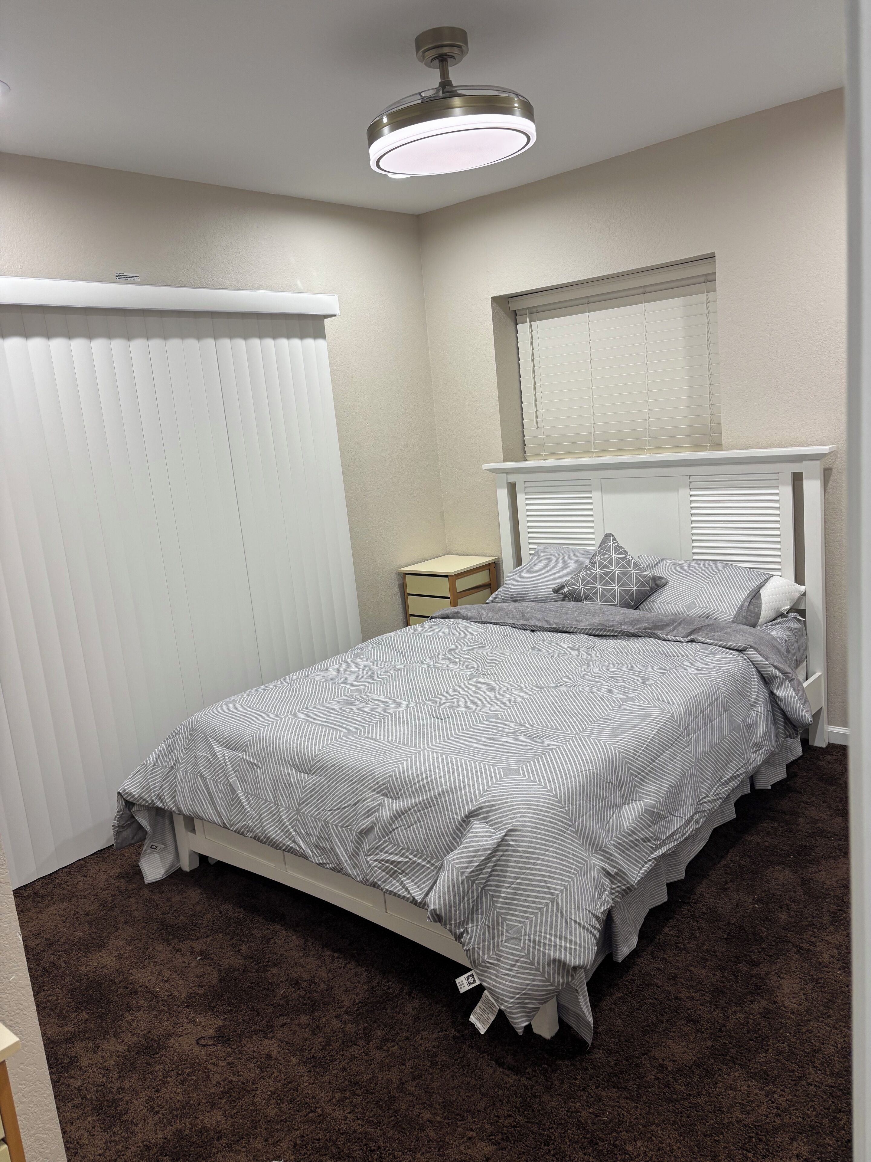 A bedroom with a bed, a nightstand, a window with blinds, and a ceiling fan.