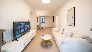 Un salon moderne avec un téléviseur à écran plat, un canapé blanc, une table basse ronde et, au fond, un coin repas.