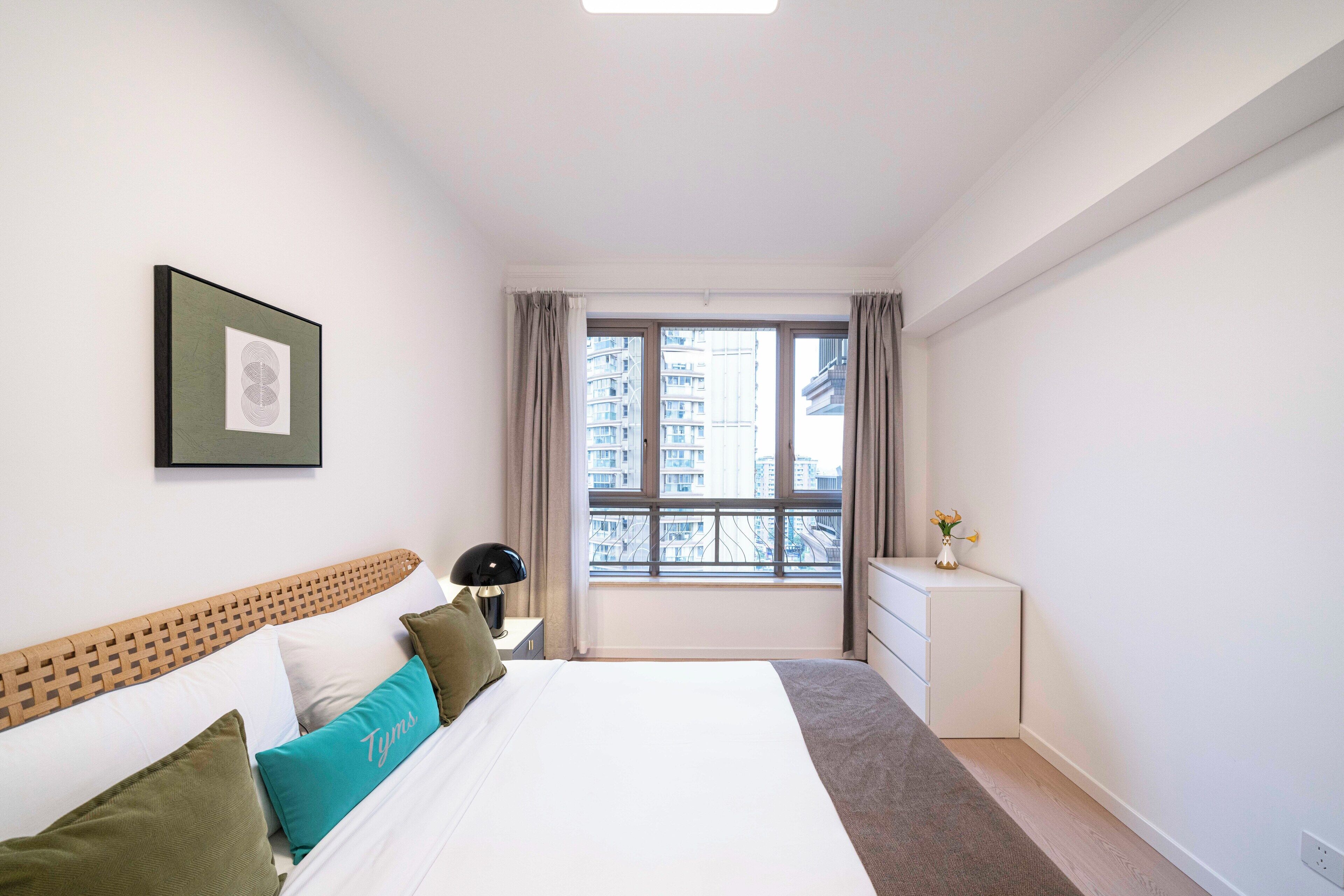A modern bedroom with a large bed, a nightstand, a dresser, and a window with a city view.