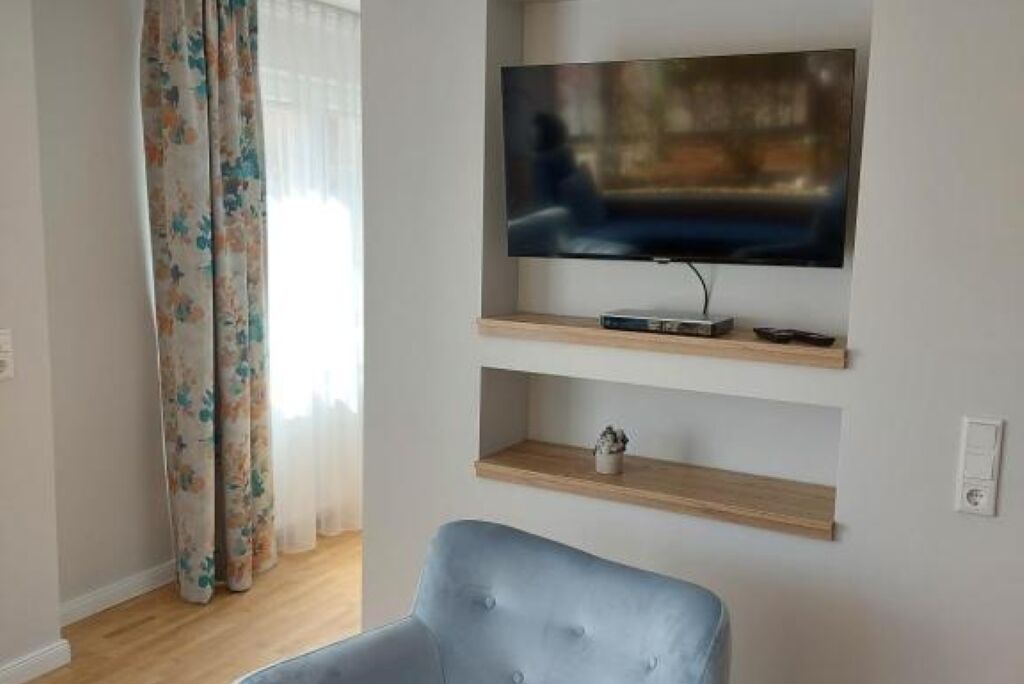 Ein Wohnzimmer mit einem an der Wand befestigten Flachbildfernseher, darunter hölzerne Regale und ein blauer Sessel.