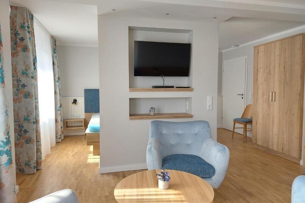 A modern living room with a flat-screen TV mounted on the wall, a blue armchair, a wooden coffee table, and a wooden wardrobe.
