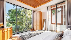 A bedroom with a large bed, wooden wardrobe, and a view of greenery through floor-to-ceiling windows. - Villa Temple Tree By Soobali - UBUD (Ubud)