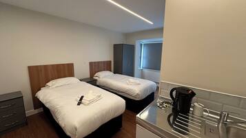 Una habitación de hotel con dos camas, un kitchenette pequeño, una cafetera y un ventanal con persianas.