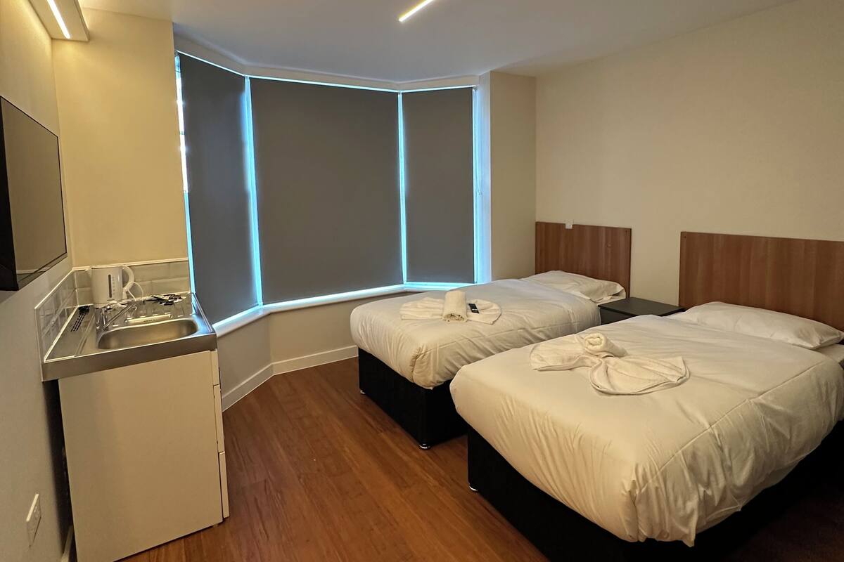 A modern hotel room with two beds, a small kitchenette, and a corner window with built-in lighting.