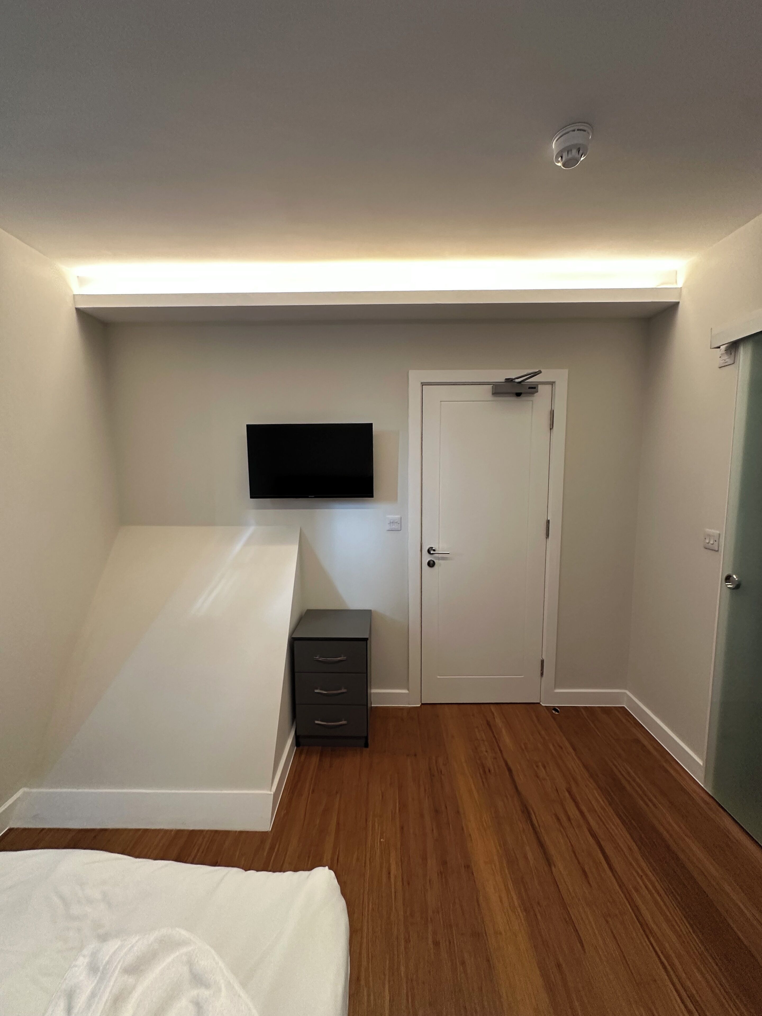 A bedroom with a bed, a flat-screen TV mounted on the wall, a door, and a small cabinet.
