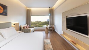 A modern hotel room with a large bed, a TV, a desk, and a view of the hills. - Regenta Hotel Science Center Bhuj (Bhuj)