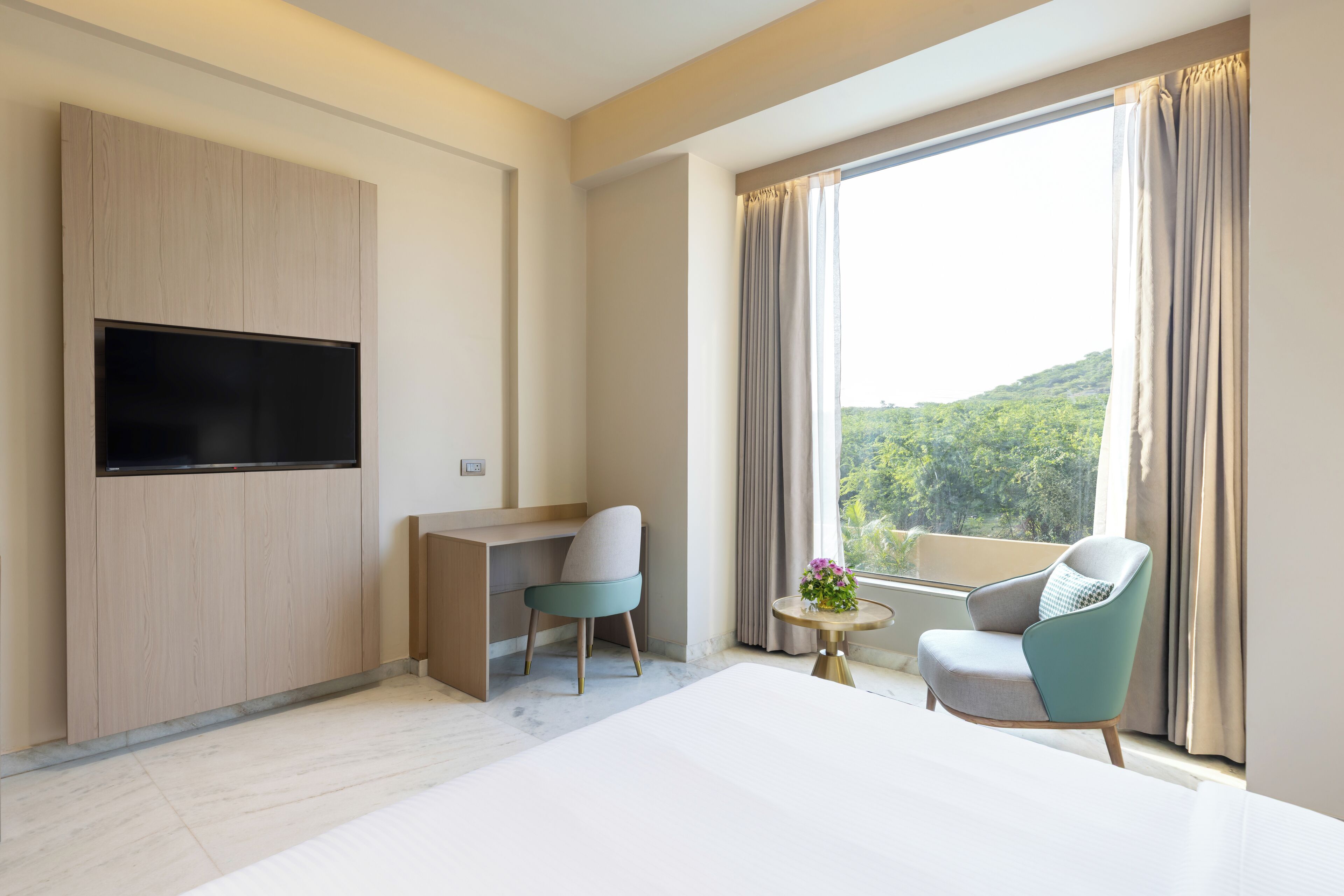 A modern hotel room with a large bed, a desk, two chairs, a TV, and a balcony with a scenic view.