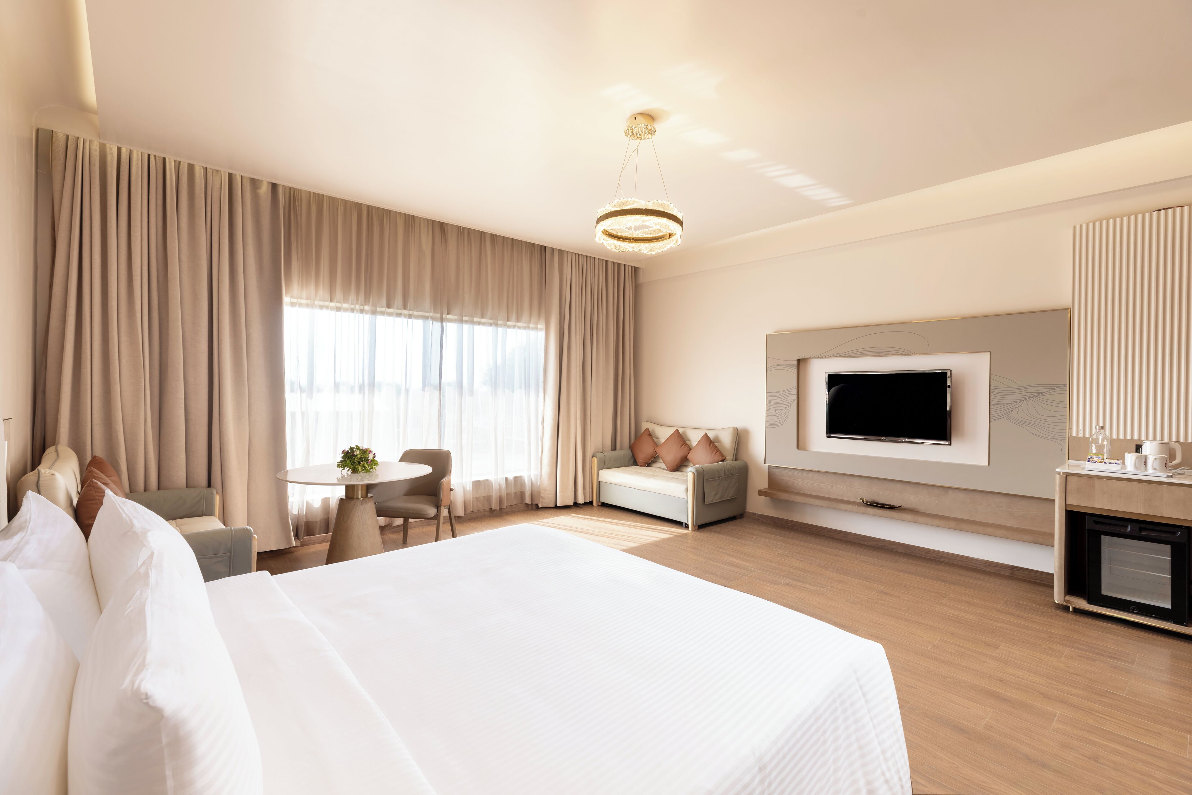 Una habitación de hotel moderna con una cama grande, un televisor montado en la pared, una zona de estar con una mesa y sillas, y una kitchenette.