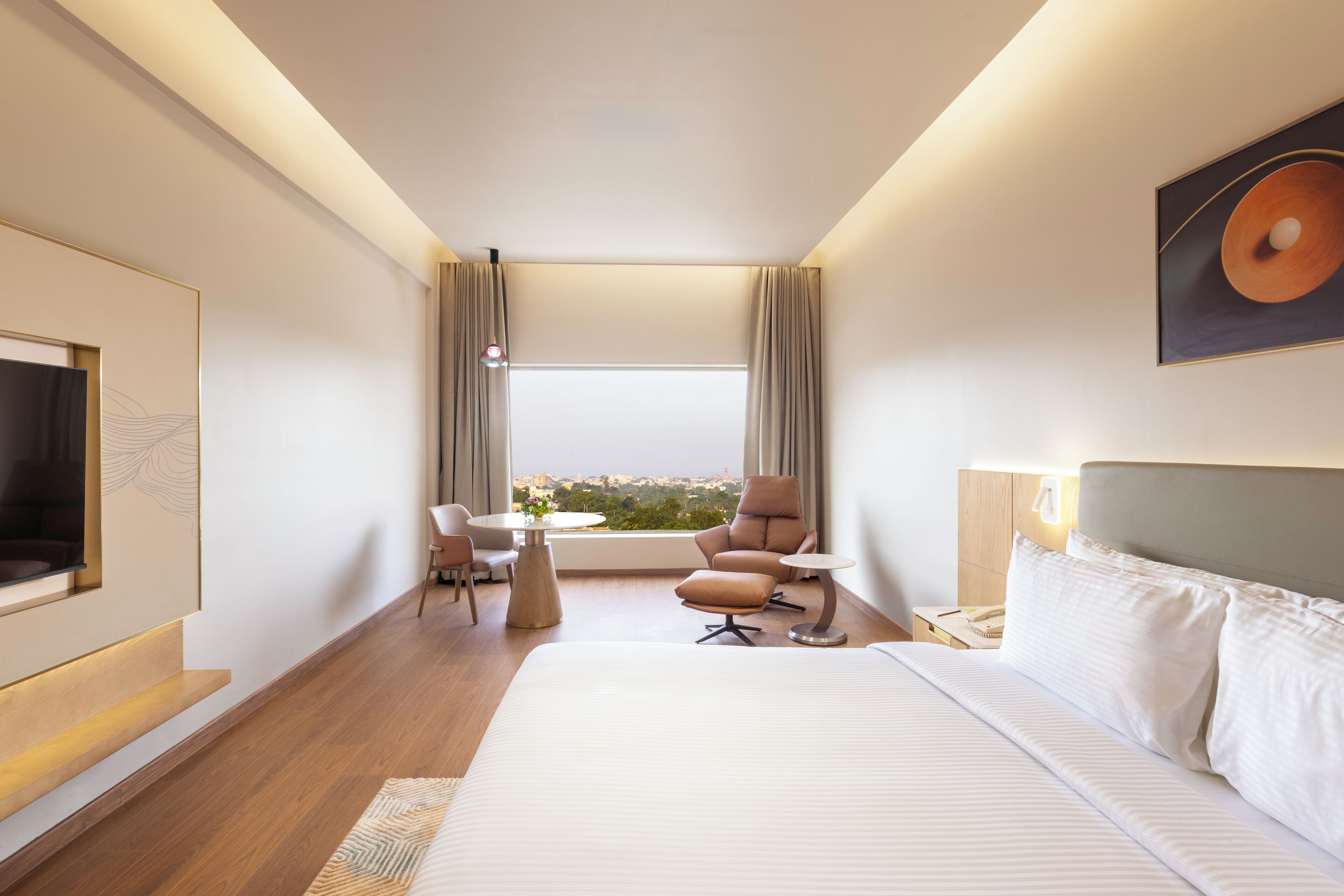 A modern hotel room with a large bed, a TV mounted on the wall, a desk with a chair, and a window offering a city view.