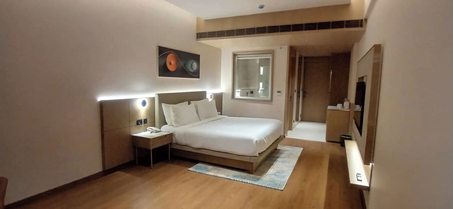A modern hotel room with a bed, bedside table, lamp, and a painting on the wall.