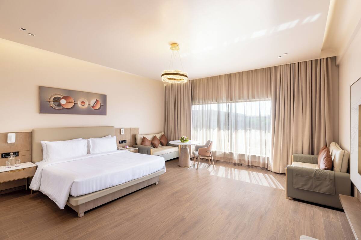 Moderni hotellihuone, jossa on suuri sänky, sohva, pieni pöytä ja tuoli. Seinällä on taulu ja ikkunassa verhot.