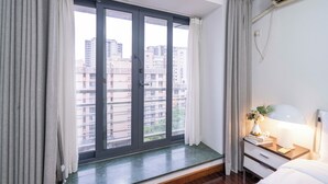 Habitación de hotel con un amplio ventanal corredero a un balcón, una mesita de noche con lámpara y vistas a la ciudad.
