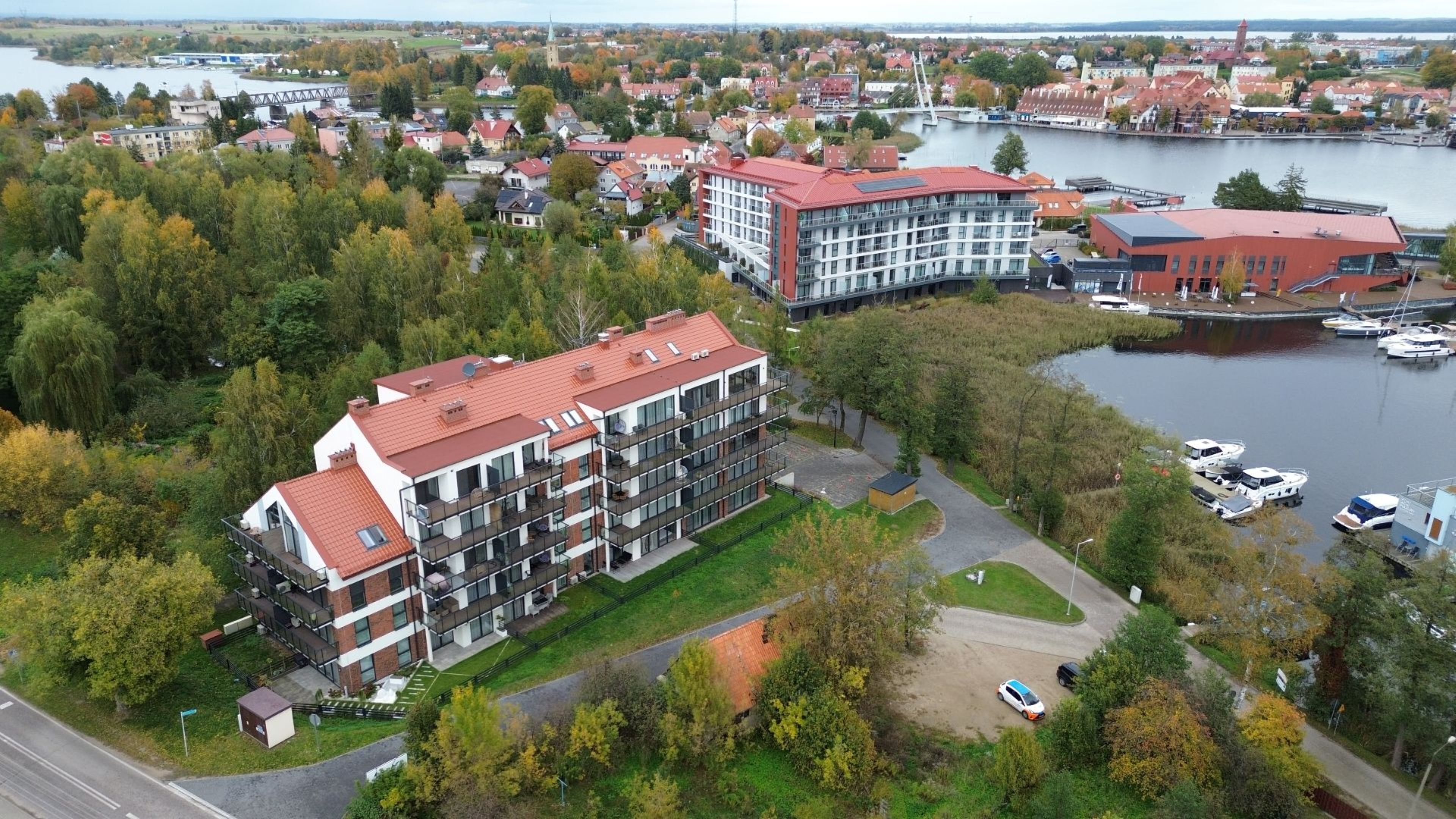 An aerial view of a modern apartment complex with multiple floors, a parking area, and a river in the background.