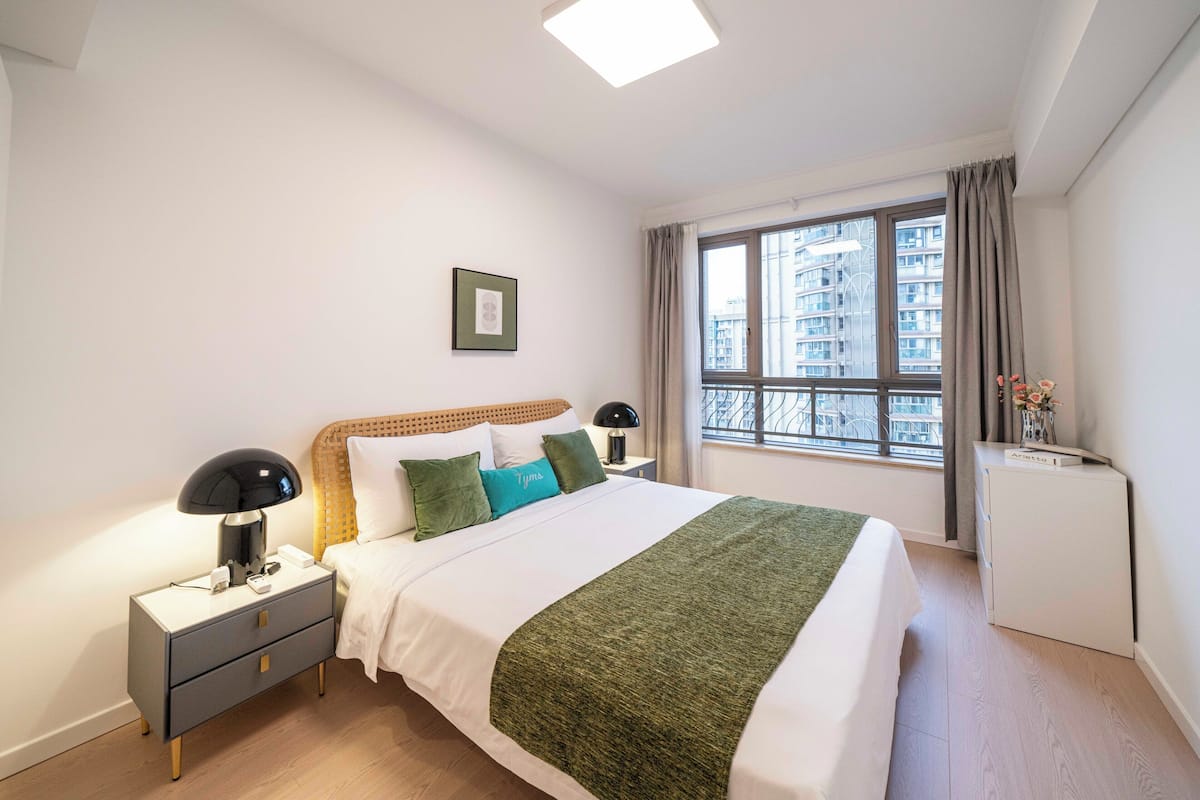 Une chambre bien rangée, avec un lit, des tables de chevet, une commode, une fenêtre avec des rideaux et une petite armoire blanche.