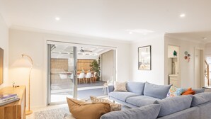 A modern living room with a grey sofa, a coffee table, and a floor lamp. There is a sliding glass door leading to a dining area with a table and chairs. - Capri Blue 2, 88 Tomaree Rd - stunning townhouse with Wi-Fi, Air Con (Shoal Bay)
