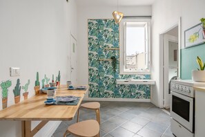A kitchen with a wooden table, two stools, a window, and a wall with a tropical leaf pattern. - 'Fiorelli 17 - Bivano Green Leaves' Apartment with Balcony, Wi-Fi and Air Conditioning (Palermo)