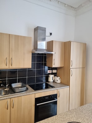 A kitchen with wooden cabinets, a black tiled backsplash, a stainless steel range hood, a black oven, a sink, and a countertop with a kettle and toaster. - Flat In Tyne & Wear Sunderland (Sunderland)