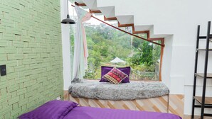 A bedroom with a view of greenery, a bed with purple bedding, a window seat with a cushion, and a staircase. - SWEET VALLEY HOMESTAY (Xuan Huong - Da Lat)