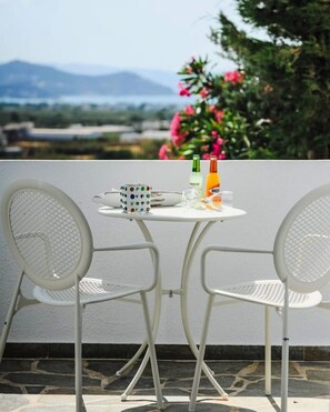 Ein weißer Metalltisch mit zwei Stühlen, einer Tasse und zwei Flaschen auf einem Balkon mit Blick auf Berge und Blumen.