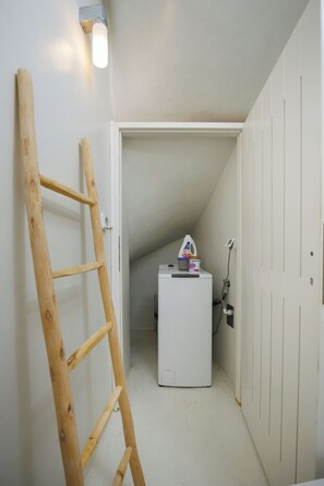 A narrow hallway with a wooden ladder leaning against the wall, a white refrigerator, and a door leading to another room.