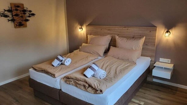 Uma cama de casal com duas almofadas e duas toalhas enroladas, uma mesinha de cabeceira e um aparelho de luz fixado na parede.