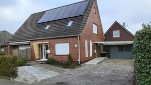 Uma casa de dois andares com telhado inclinado, painéis solares, garagem e um pequeno jardim frontal.