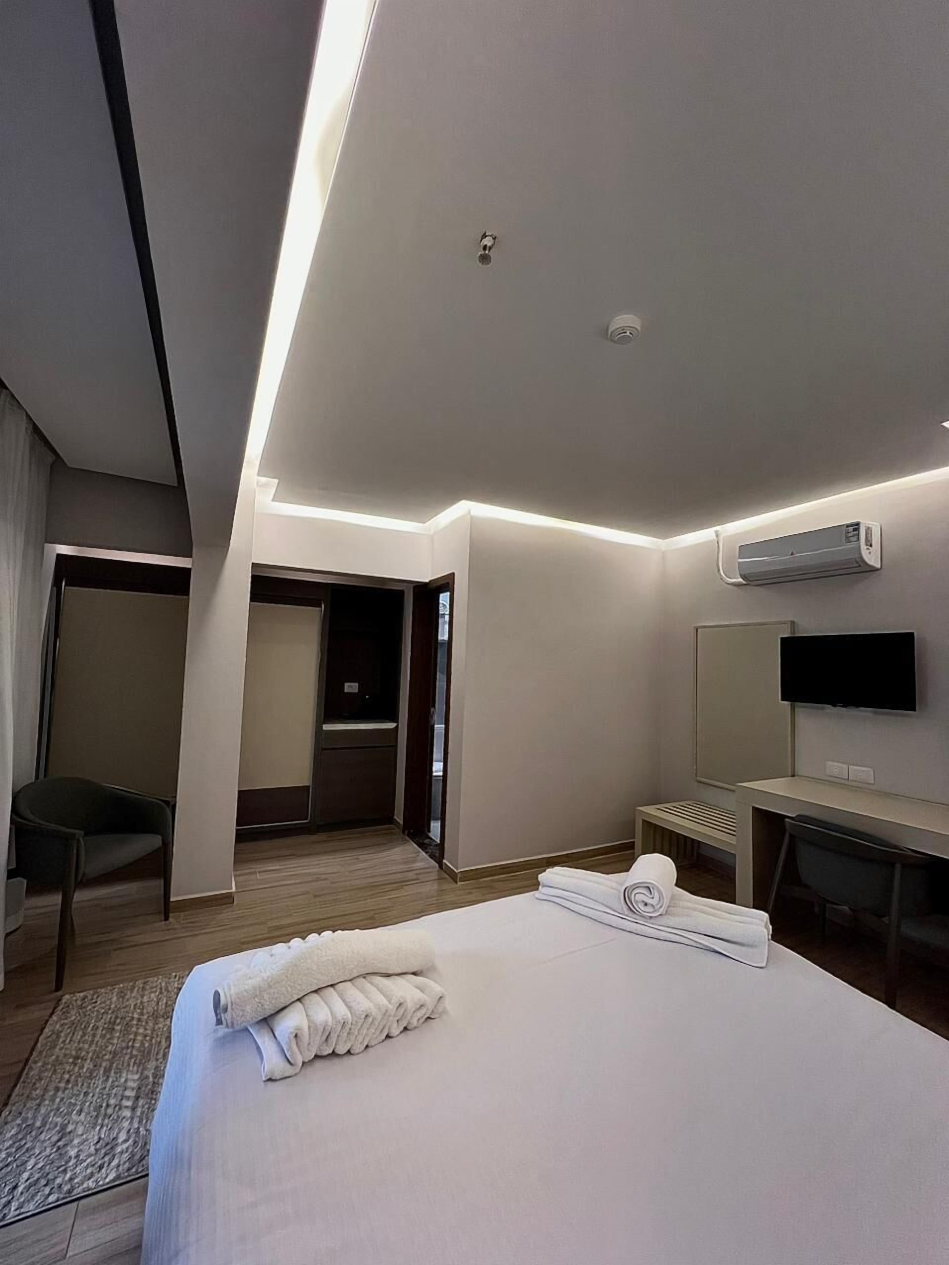 A modern hotel room with a neatly made bed, a desk with a chair, a flat-screen TV, and an air conditioning unit.