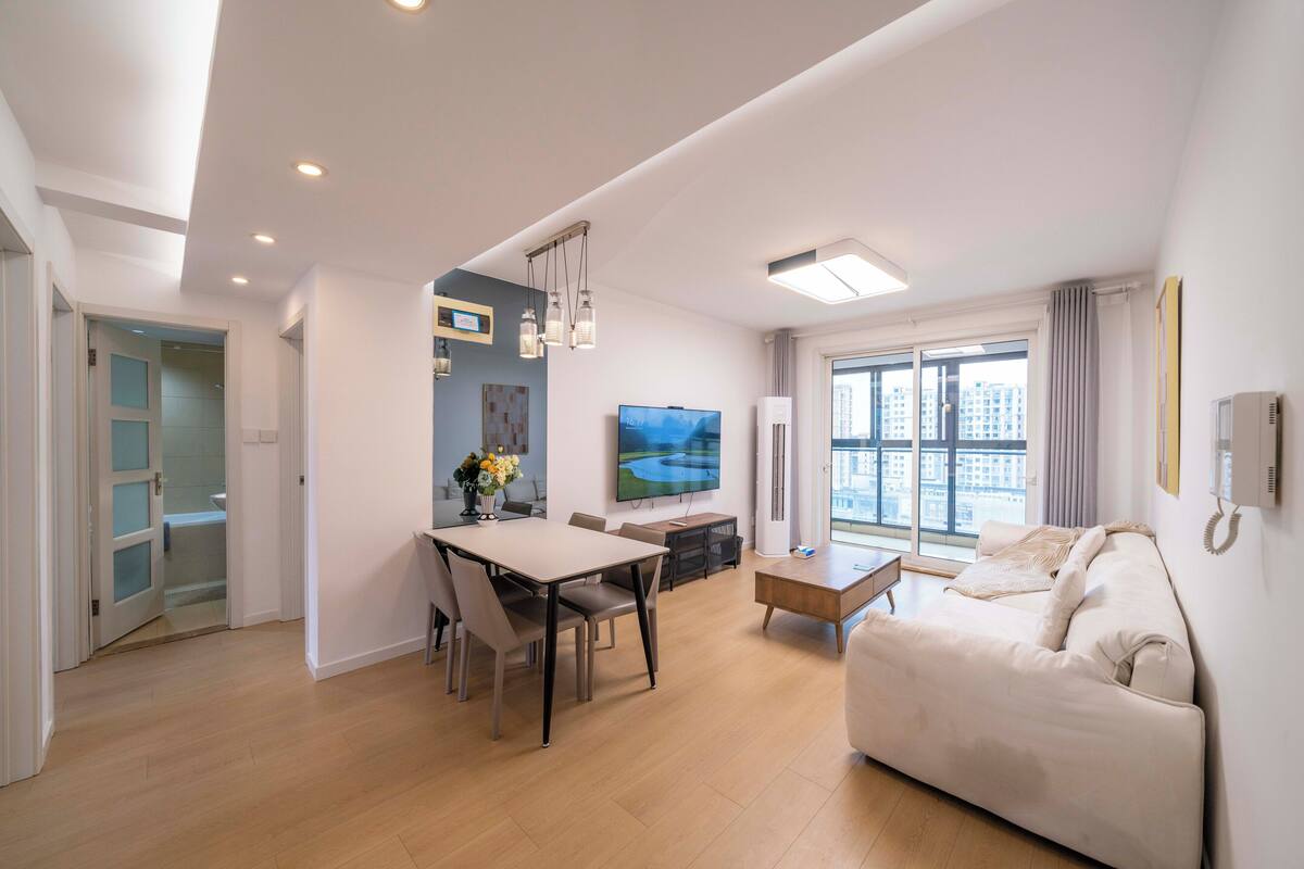 A modern living room with a sofa, a dining table with chairs, a TV, and a large window with curtains.