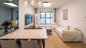 A modern living room with a white sofa, a dining table with chairs, a TV mounted on the wall, and a large window with a city view. - Tyms Family APT Spacious 3BR for Groups & Families  (Shanghai)