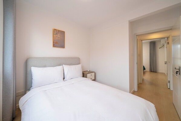 A hotel room with a bed, a nightstand, a painting on the wall, and a hallway leading to another room. - Tyms Family APT Spacious 3BR for Groups & Families (Shanghai)
