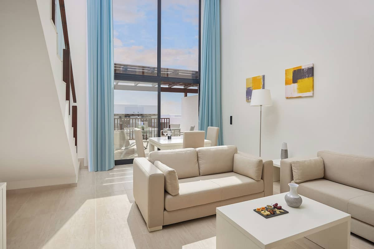A modern living room with a beige sofa, a white coffee table with a fruit platter, and a floor lamp. There is a large window with blue curtains and a view of a balcony.