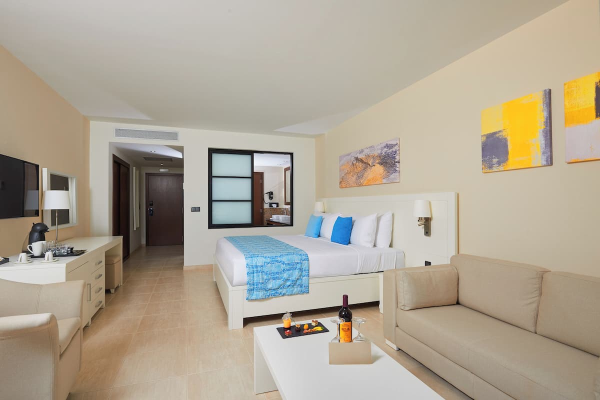 A hotel room with a bed, a sofa, a desk, and a coffee table. There are paintings on the wall and a door leading to another room.