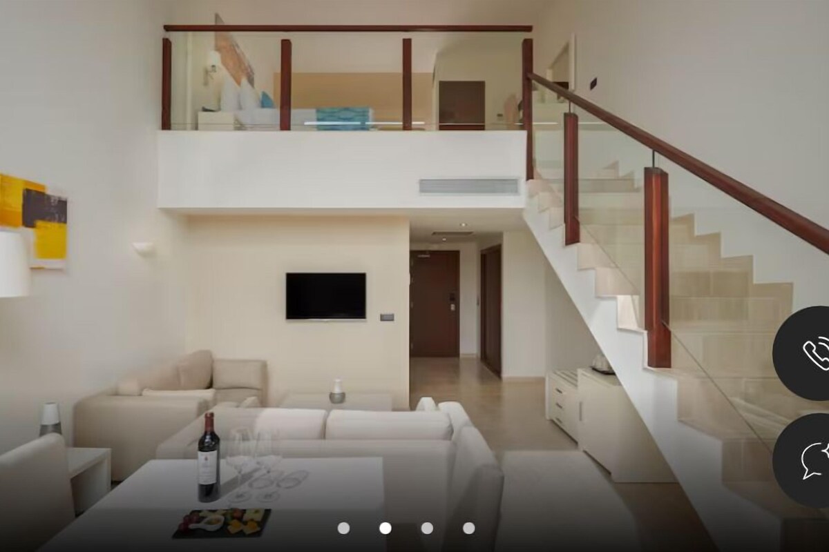 Un salon moderne avec un ensemble canapé, une table basse sur laquelle se trouve une bouteille, et un téléviseur à écran plat fixé au mur.
